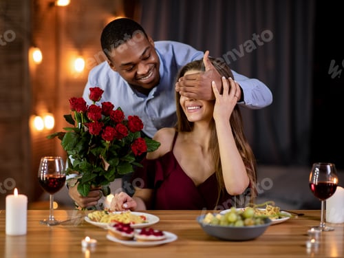 Preview: Romantic Surprise. Happy black man covering girlfriend's eyes and giving roses bouquet