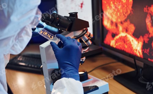 Preview: Scientist in white protective wear indoors in laboratory with lens with virus title on it