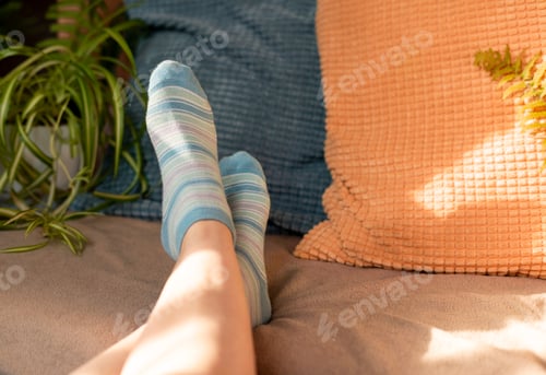 Preview: Female legs in blue striped socks on sofa among pillows against background of potted plants resting