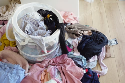 Preview: Clothes pile next to laundry basket filled with garments, on hardwood floor
