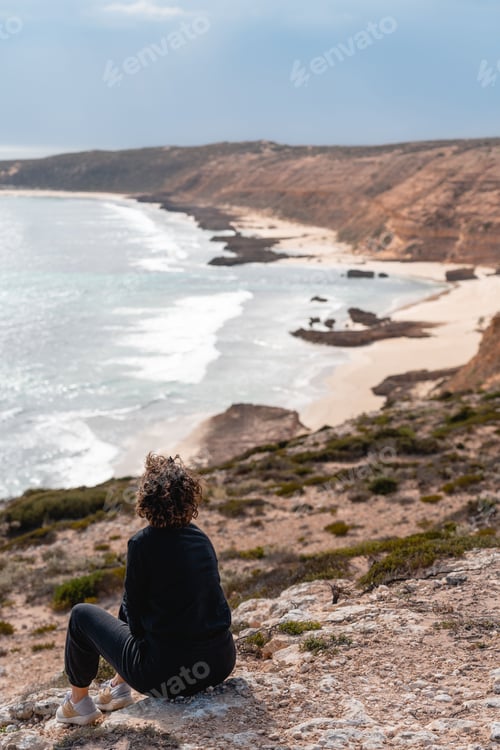 Preview: Woman with short hair sitting on a rock at the beach and looking to the sea in a magical landscape