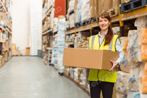 Preview: Female worker holding cardboard box standing in warehouse aisle with vest and shelves, copy space