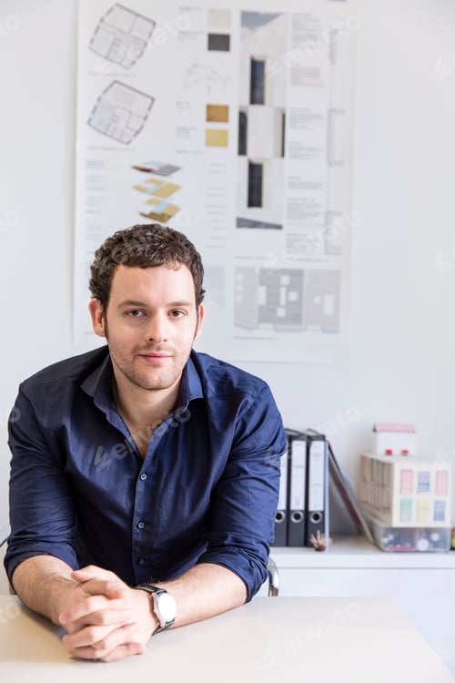 Preview: Mid adult man at desk in office in front of blueprint looking at camera