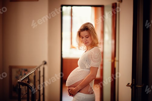 Preview: Blonde pregnant woman in white knitted clothes hold her big belly with smile on her face