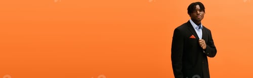 Preview: Handsome young man in stylish black suit poses confidently against a vivid orange background