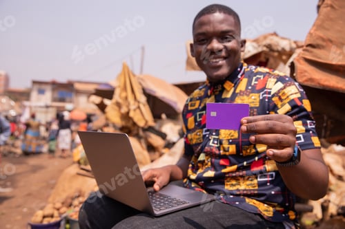 Preview: African boy shows his credit card while using his laptop, he is at the market. Credit card in Africa