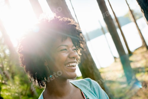 Aperçu: A young woman with curly black hair in the shade of trees by a lake shore.