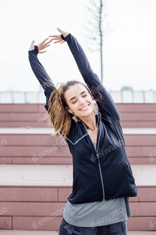 Preview: Fit young woman with earphones doing stretching exercises on stairs outdoors