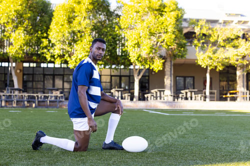Preview: African American young male athlete kneeling on field outdoors, holding a rugby ball, copy space