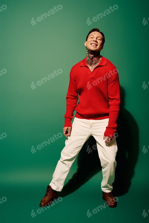 Preview: Young man with tattoos smiling joyfully against a green backdrop in a stylish red sweater
