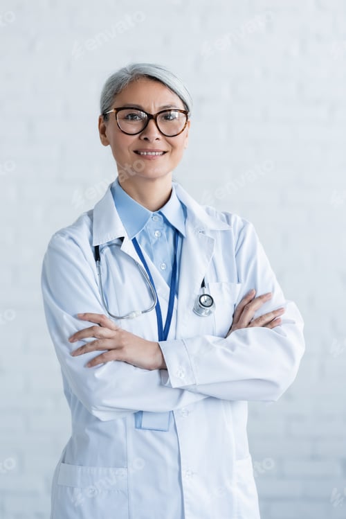 Preview: mature asian doctor in white coat and eyeglasses standing with crossed arms and smiling at camera on