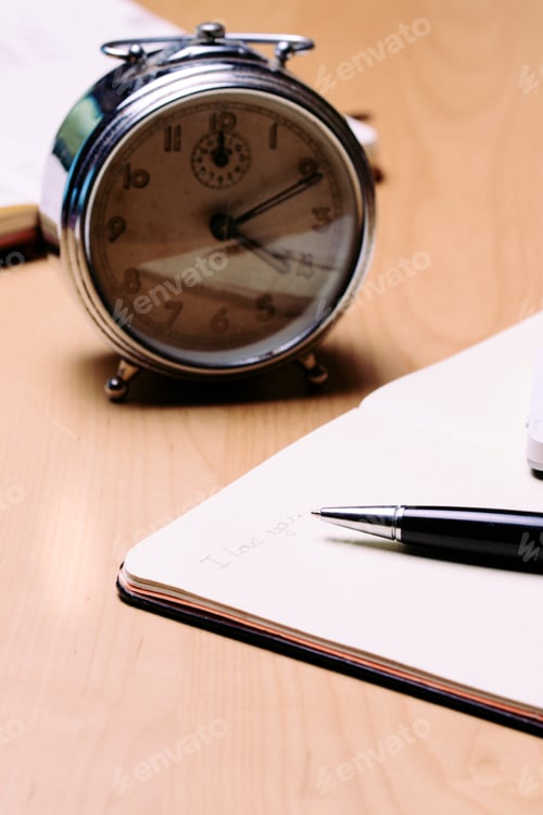 Preview: Note book with an alarm clock in the background.