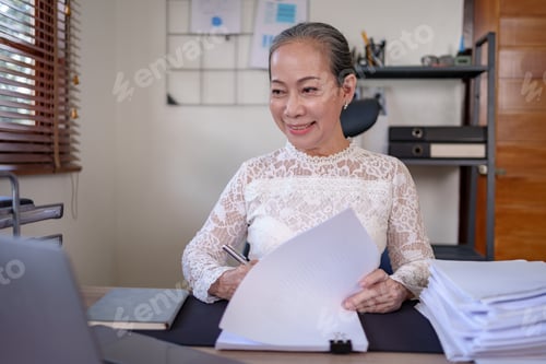 Preview: Senior asian business woman sitting checking paperwork on a desk with a laptop in the office.