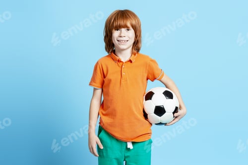 Preview: Redhead boy enjoying soccer, smiling and holding a football on blue background