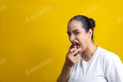 Preview: Hispanic woman biting a red plum, on yellow background