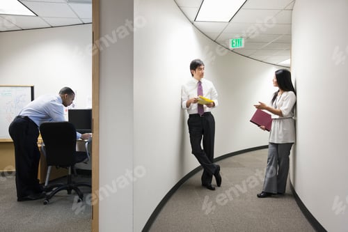 Preview: Two asian businesspeople talking in a hallway near a black businessman working alone in his office.