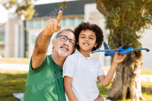 Preview: Positive senior man with beard and black little boy hug, have fun in park, flying toy airplane