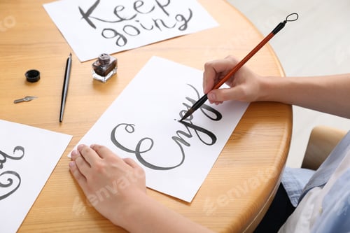 Preview: Calligraphy. Woman with brush writing word Enjoy on sheet of paper at wooden table indoors, closeup