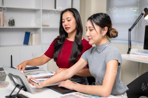 Preview: Pretty asian woman and coworker looking at tablet aside working materials on table in the office.