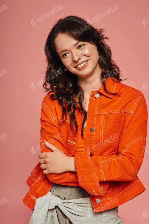 Preview: Young woman radiates joy in vibrant orange jacket against a playful pink backdrop