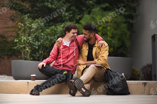 Preview: Happy young man with Down syndrome and mentoring friend sittingwith arms around outdoors
