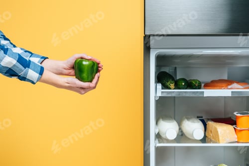 Preview: cropped view of woman holding bell pepper near open fridge with fresh food on shelves isolated on