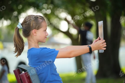 Preview: Young cute smiling teenager girl looking at screen of digital tablet sitting on bench in park