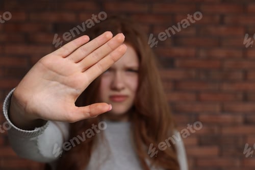 Preview: Sad teenage girl making stop gesture near brick wall indoors, selective focus