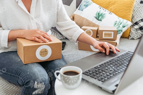 Preview: Young woman sitting on couch, working on laptop and holding box.