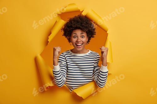 Preview: Finally, new achievement! Brunette curly adult woman in casual striped jumper makes victory gesture