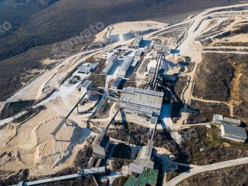 Preview: A large industrial cement plant surrounded by barren hills and dirt roads. Conveyor belts and