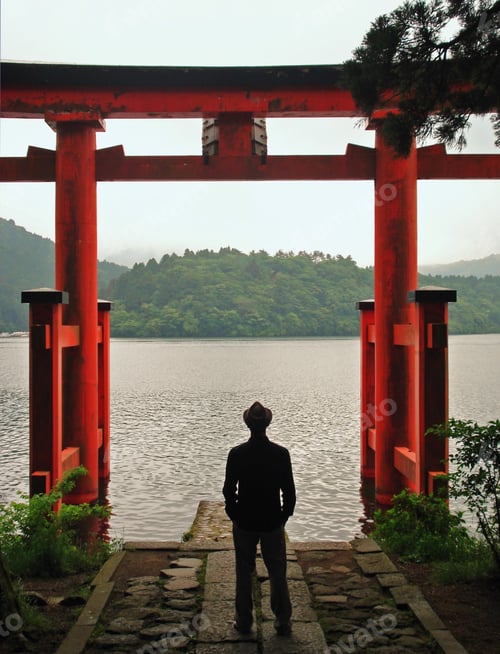 Preview: Back view of man standing beside red torii on shoreline of lake