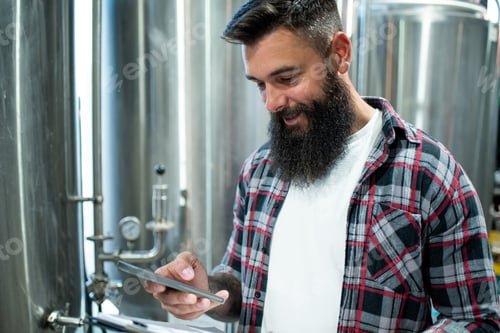 Preview: Man working in brewery and taking notes of the beer production and using smartphone