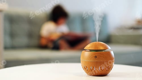 Preview: Modern aroma oil diffuser on the white table. Little boy in the background reading a book