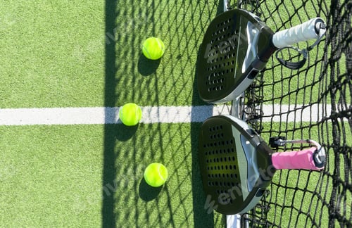 Preview: Padel racket and yellow ball behind net on a green court grass turf outdoors. Paddle is a racquet
