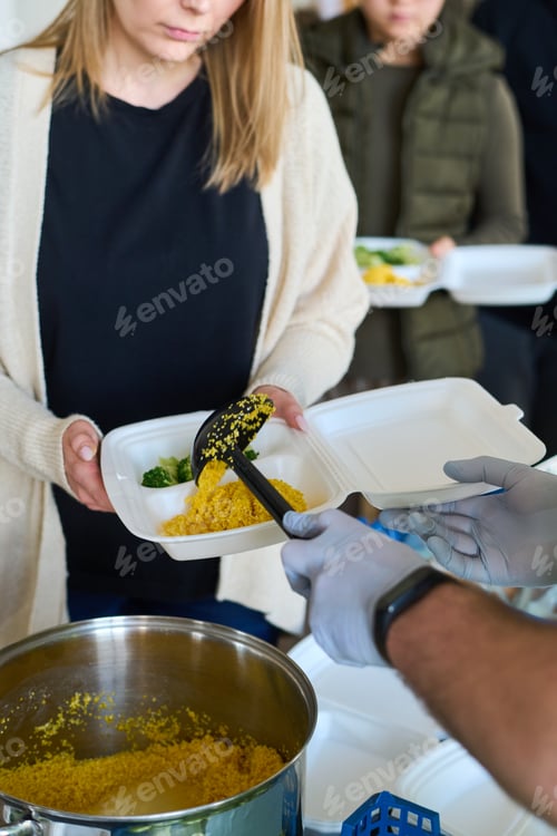 Preview: Young blond female refugee holding plastic container over pan with pilaf