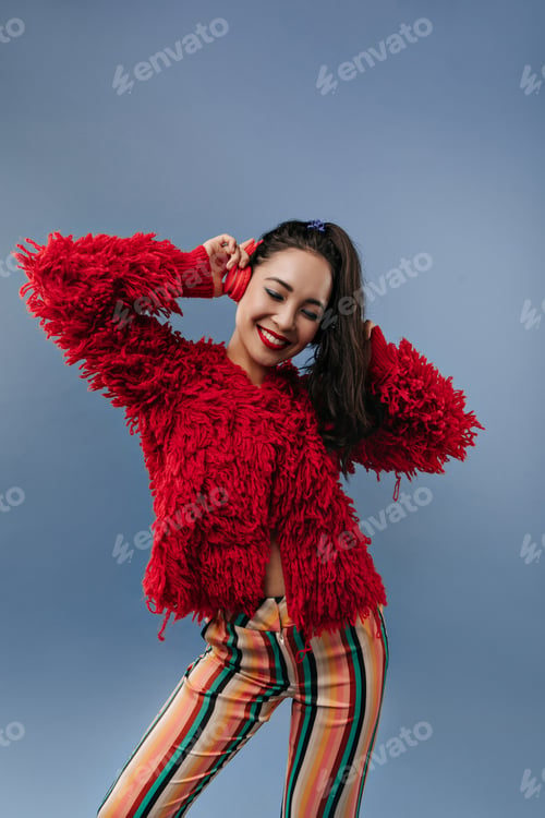 Preview: Stylish trendy woman with black hair and bright lips in warm modern sweater and striped trousers li