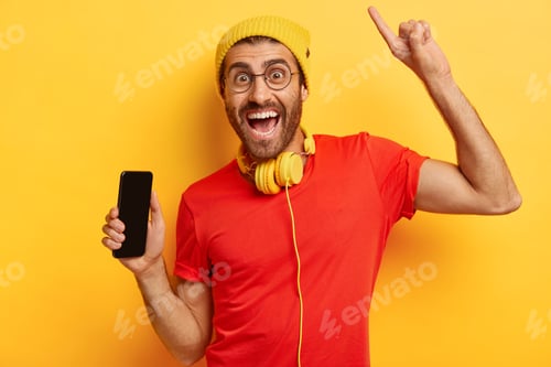 Preview: Portrait of happy unshaven man holds mobile phone with empty screen, raises arm and points with inde
