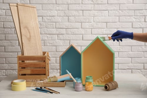 Preview: Decorator painting house shaped shelf with brush at white table, closeup. Interior element