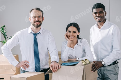 Preview: happy multiracial colleagues smiling at camera and unpacking boxes in new office