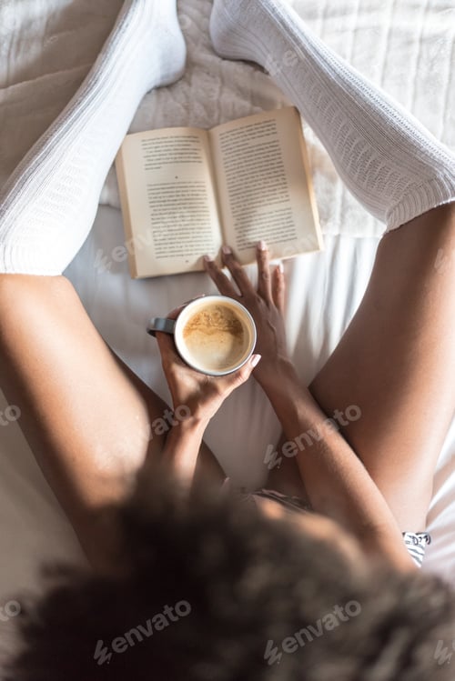 Preview: black woman reading a book and drinking coffee on bed with socks