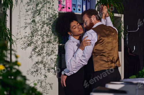 Preview: passionate interracial couple embracing near decorative plants in office at night, work romance