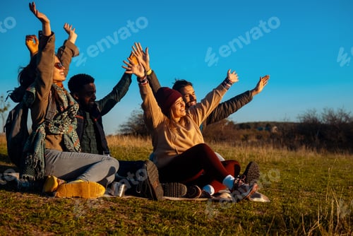 Preview: Group of friends sitting on the top of the hill resting after hiking and looking towards the sunset