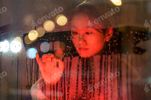 Preview: Upset asian girl suffer from solitude alone draw on wet glass in rain after breakup with boyfriend