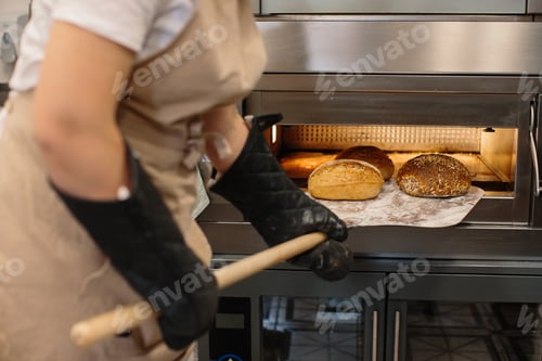 Preview: Female baker using a peel to take out a loaf of bread of the oven in a bakery