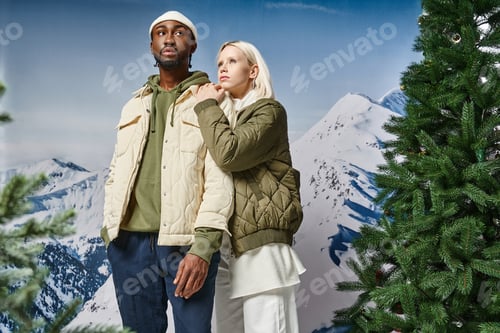 Preview: beautiful blonde woman in warm jacket hugging her stylish boyfriend in beanie hat, diverse couple