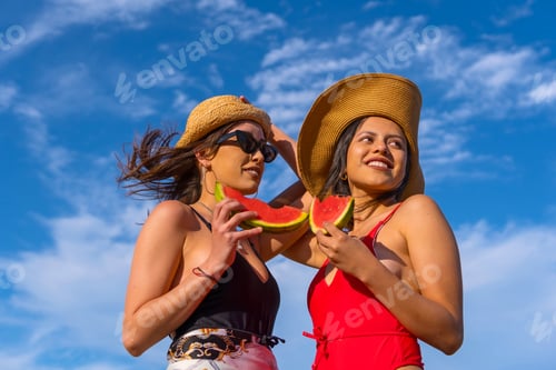 Preview: Female friends at the beach in summer smiling eating a watermelon with the sky in the background