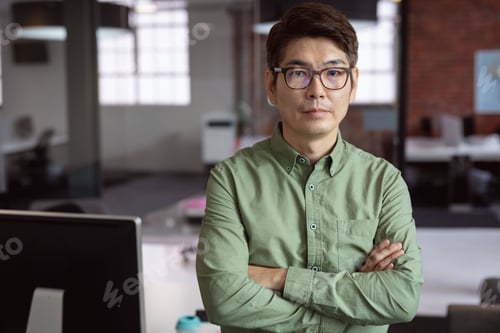 Preview: Mature Asian man standing with arms crossed in open-plan office, with monitor and water bottle