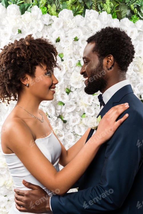 Preview: happy african american bride hugging handsome bearded bridegroom near flowers
