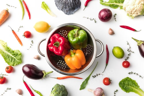 Preview: Top view of colander with colored bell peppers among uncooked vegetables isolated on white